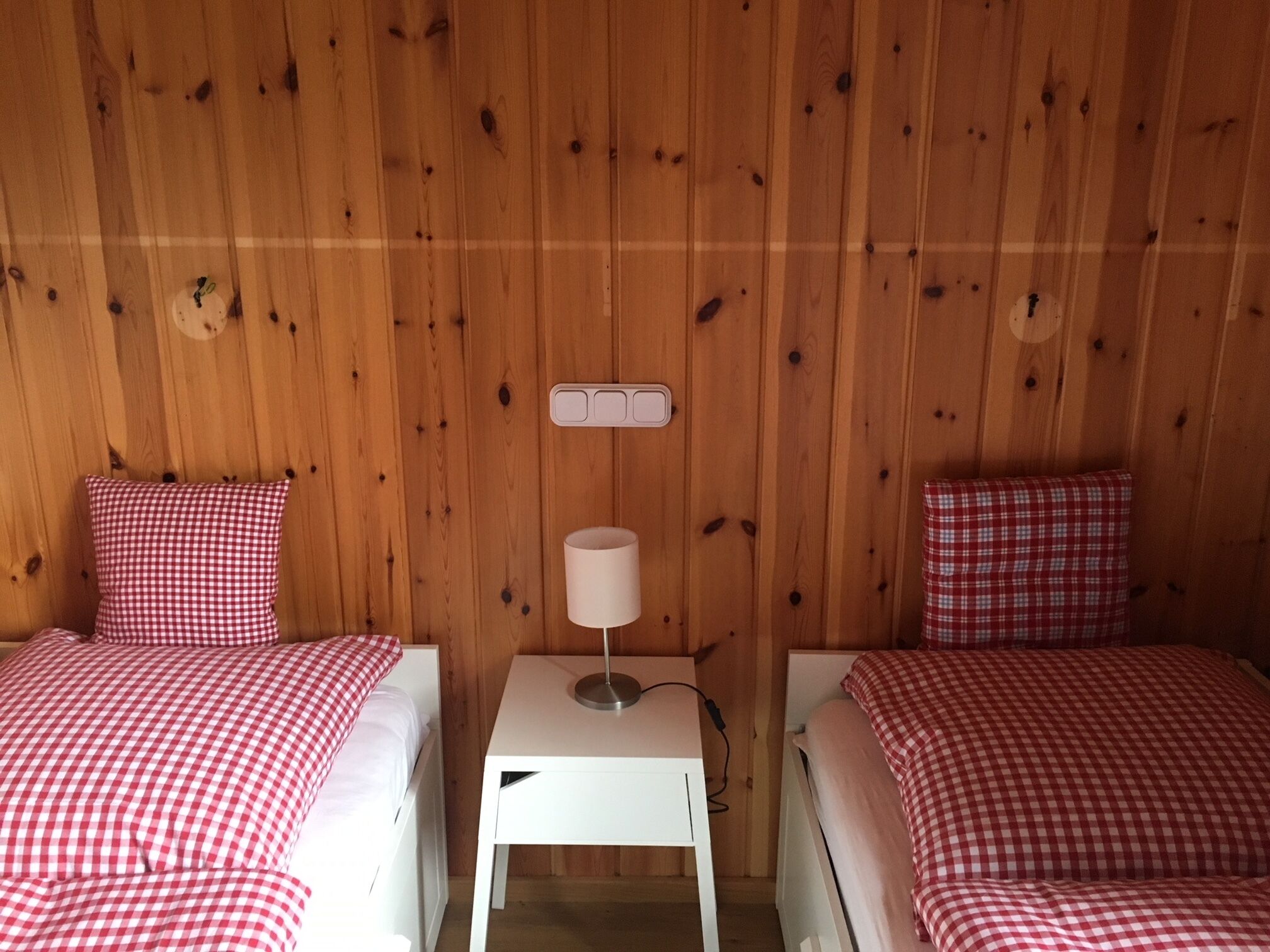 Une chambre avec deux lits, chacun recouvert d’une couverture à carreaux rouges et blancs, une table de chevet blanche avec une lampe, et un mur en bois muni d’une prise de courant.