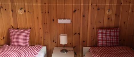 Une chambre avec deux lits, chacun recouvert d’une couverture à carreaux rouges et blancs, une table de chevet blanche avec une lampe, et un mur en bois muni d’une prise de courant.