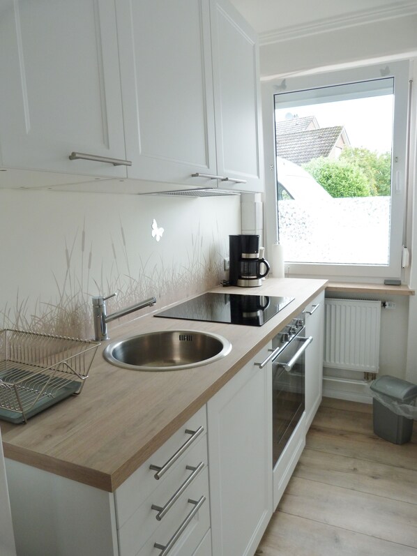A modern kitchen with white cabinets, a stainless steel sink, a black coffee maker, and a window with a view of a house and greenery. - Vacation apartment in top location, terrace, WiFi, lockable bicycle shed (Grömitz)