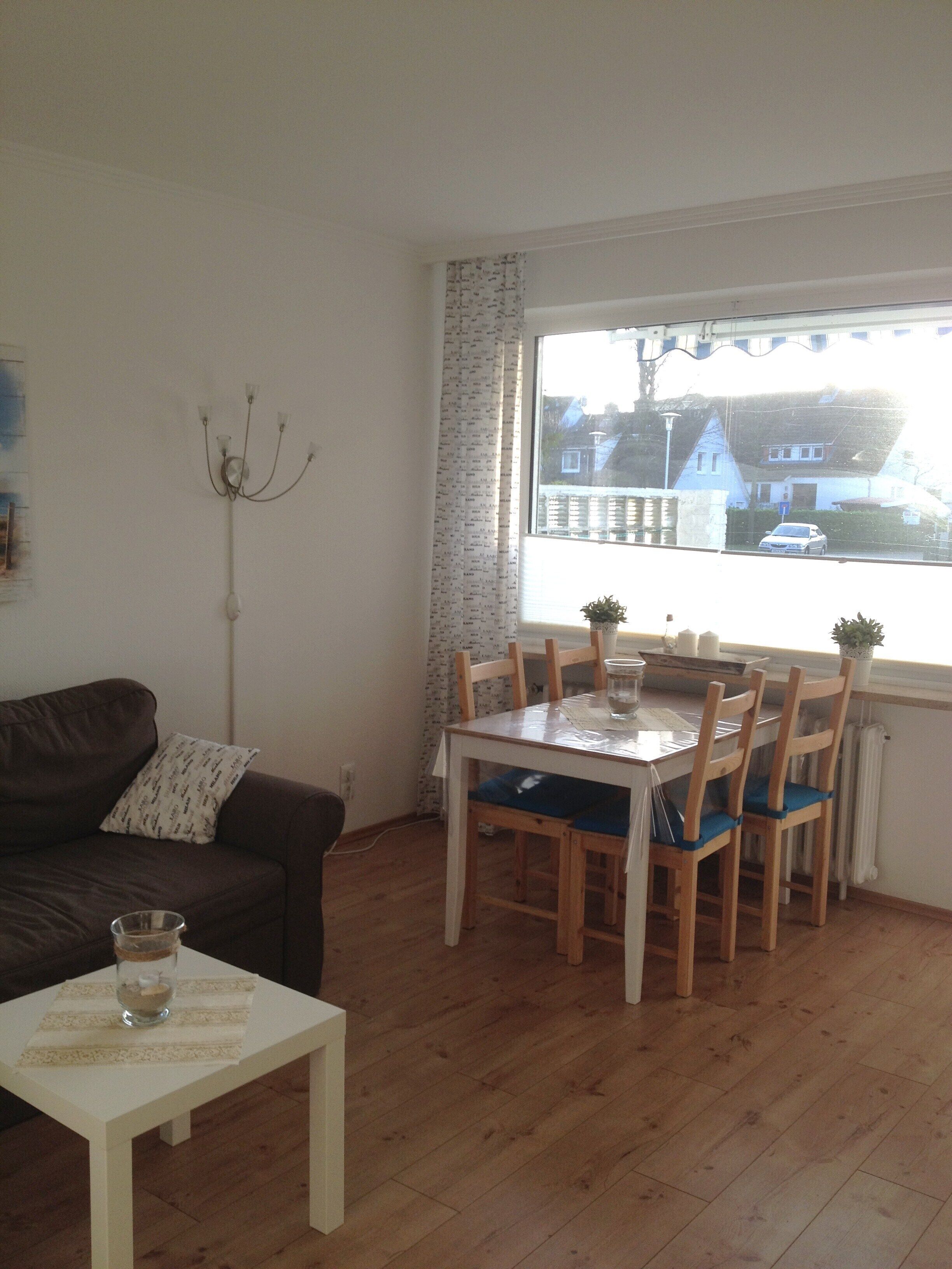 A living room with a brown sofa, a white coffee table, a dining table with four chairs, and a window with a view of houses and a car.