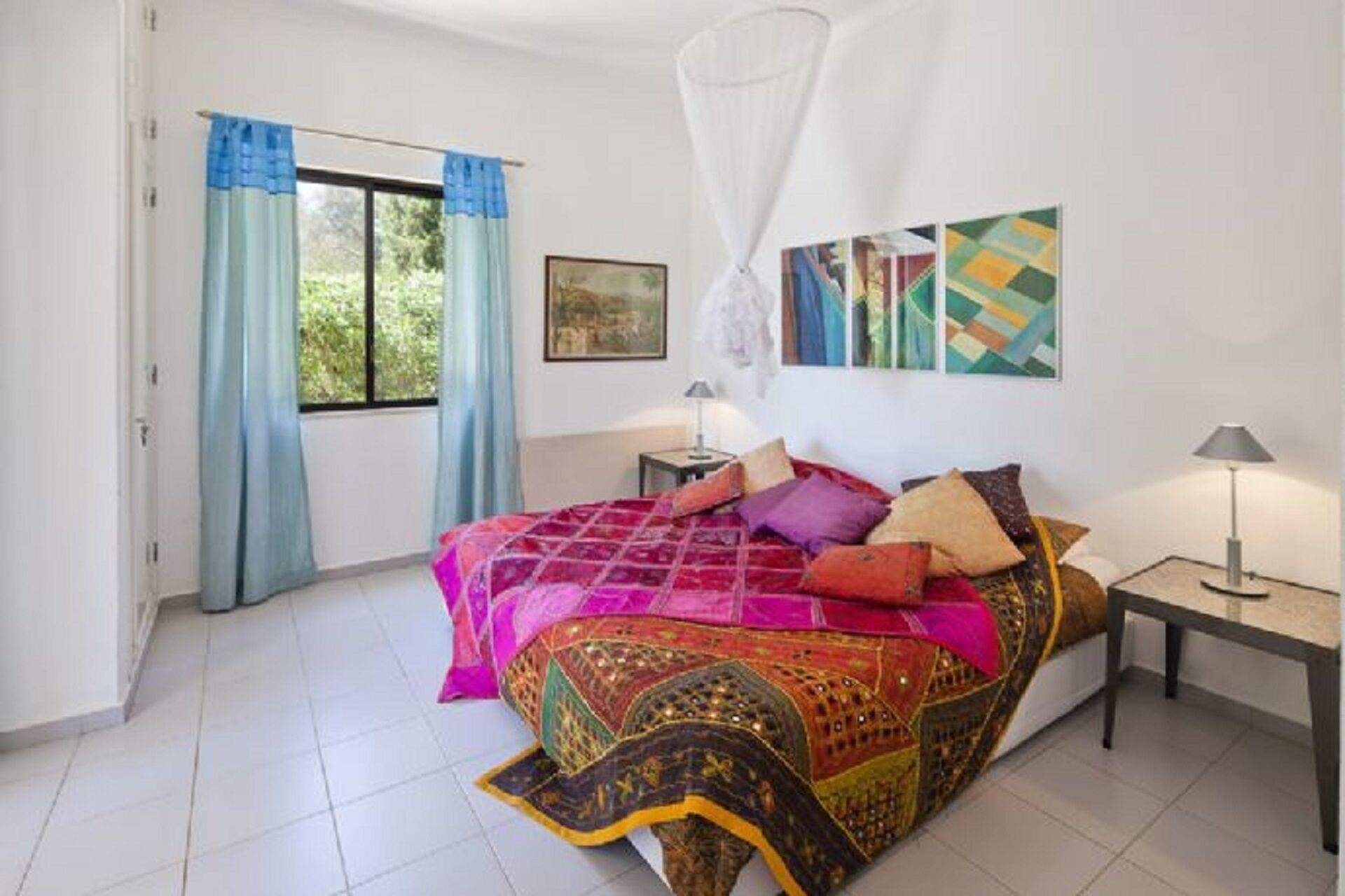 A bedroom with a bed, a bedside table, a lamp, a window with curtains, and artwork on the wall.