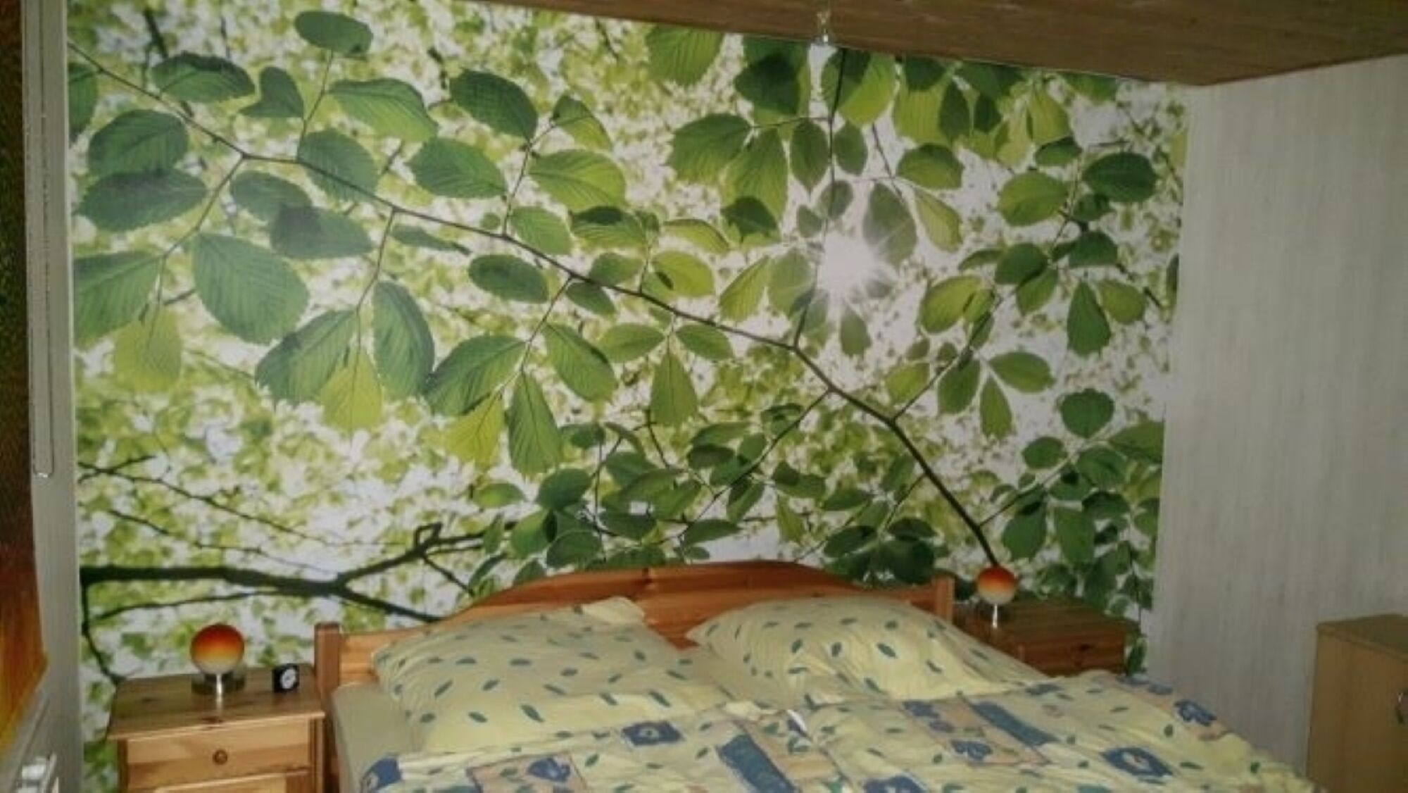 A bedroom with a bed, wooden nightstands, and a wall mural of leaves.