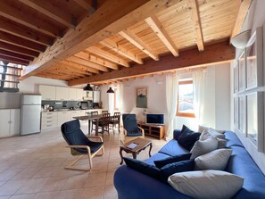 Ein geräumiger Wohnbereich mit Holzbalkendecke, einem blauen Sofa mit Kissen, einem Esstisch mit Stühlen, einer Küche im Hintergrund und einem Fenster mit Vorhängen.