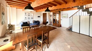 Een ruime woonkamer met houten plafond, een eettafel met stoelen, een blauwe bank en een trap naar een verdieping erboven.
