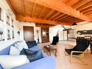 Ein großzügiger Wohnbereich mit Holzbalkendecke, einer Couch mit blauen und weißen Kissen, einem schwarzen Sessel, einem Holztisch und einer Küche im Hintergrund.