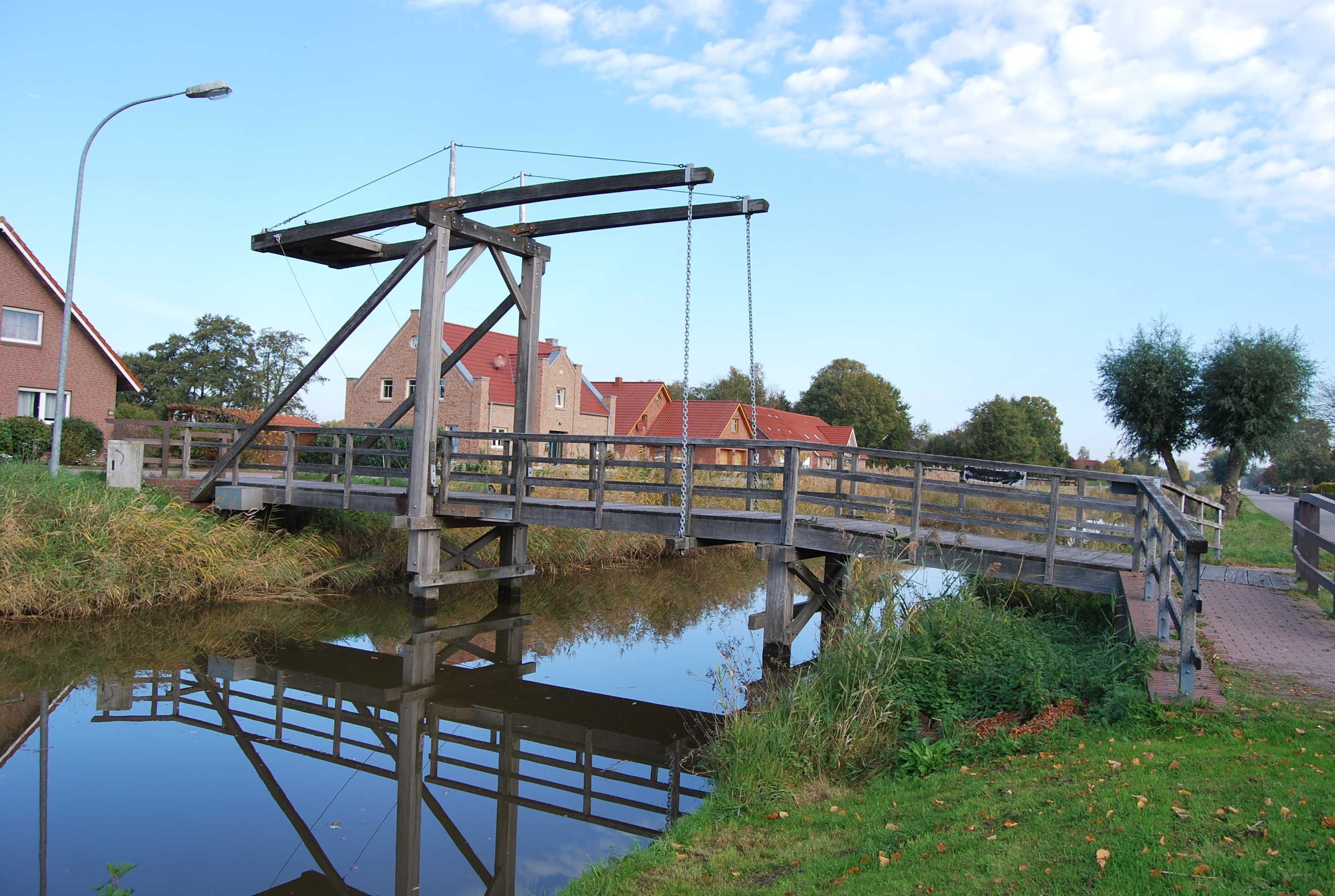 Eine Holzbrücke über ruhigem Gewässer mit Spiegelung, umgeben von Grünflächen und Wohnhäusern.