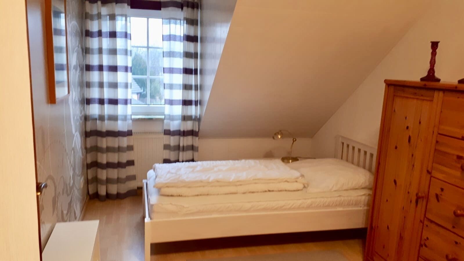 A bedroom with a bed, a wooden dresser, a window with curtains, and a radiator.