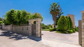 Une entrée fermée par un portail, avec des murs en pierre, une allée pavée et un aménagement paysager bien entretenu comprenant des arbres et des arbustes.