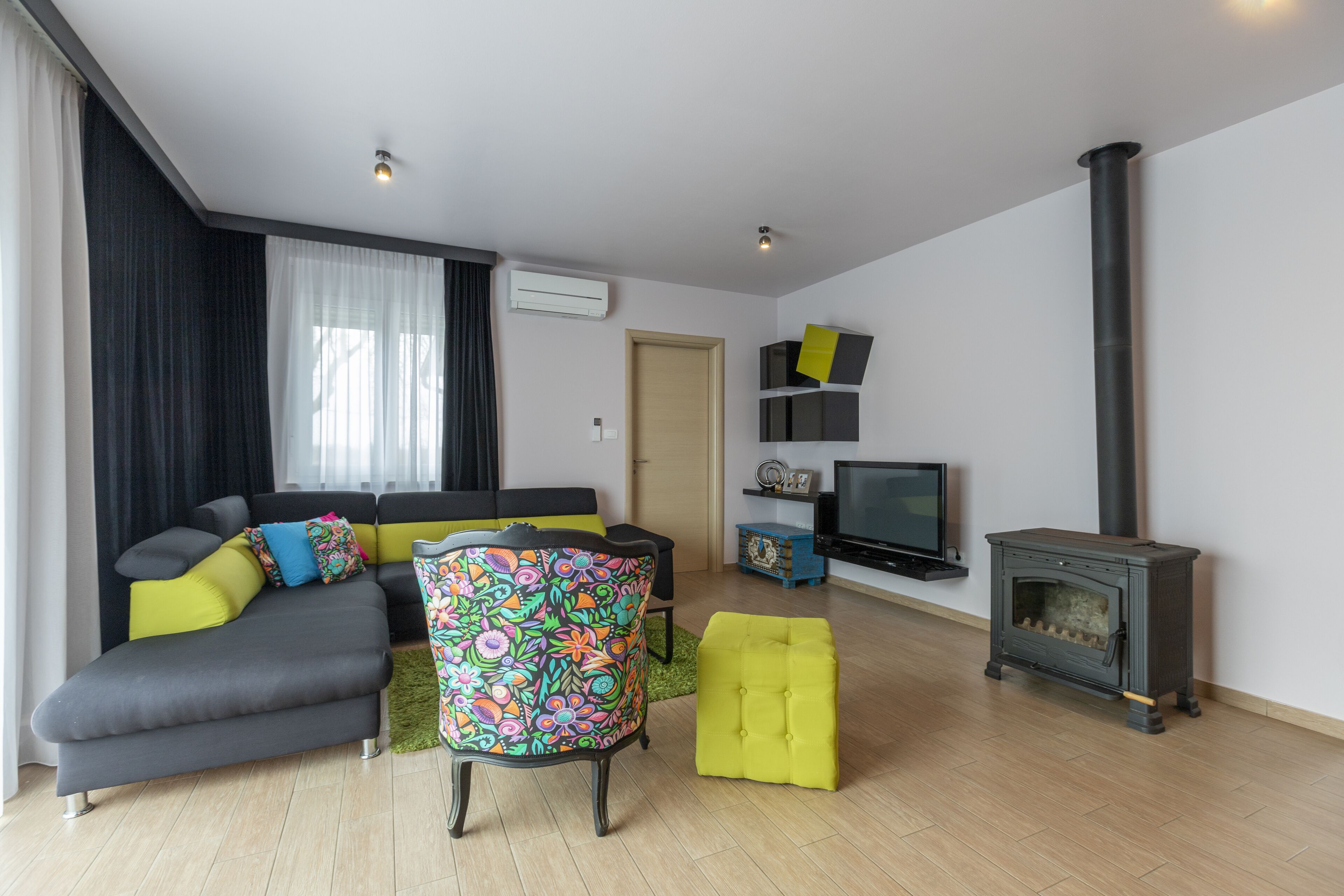 A modern living room with a sofa, colorful chair, ottoman, TV, and a wood-burning stove.
