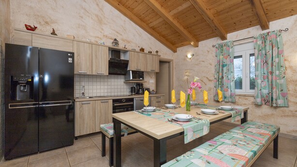 Una cocina con armarios de madera, un refrigerador negro, una mesa de comedor con bancos y una ventana con cortinas estampadas.