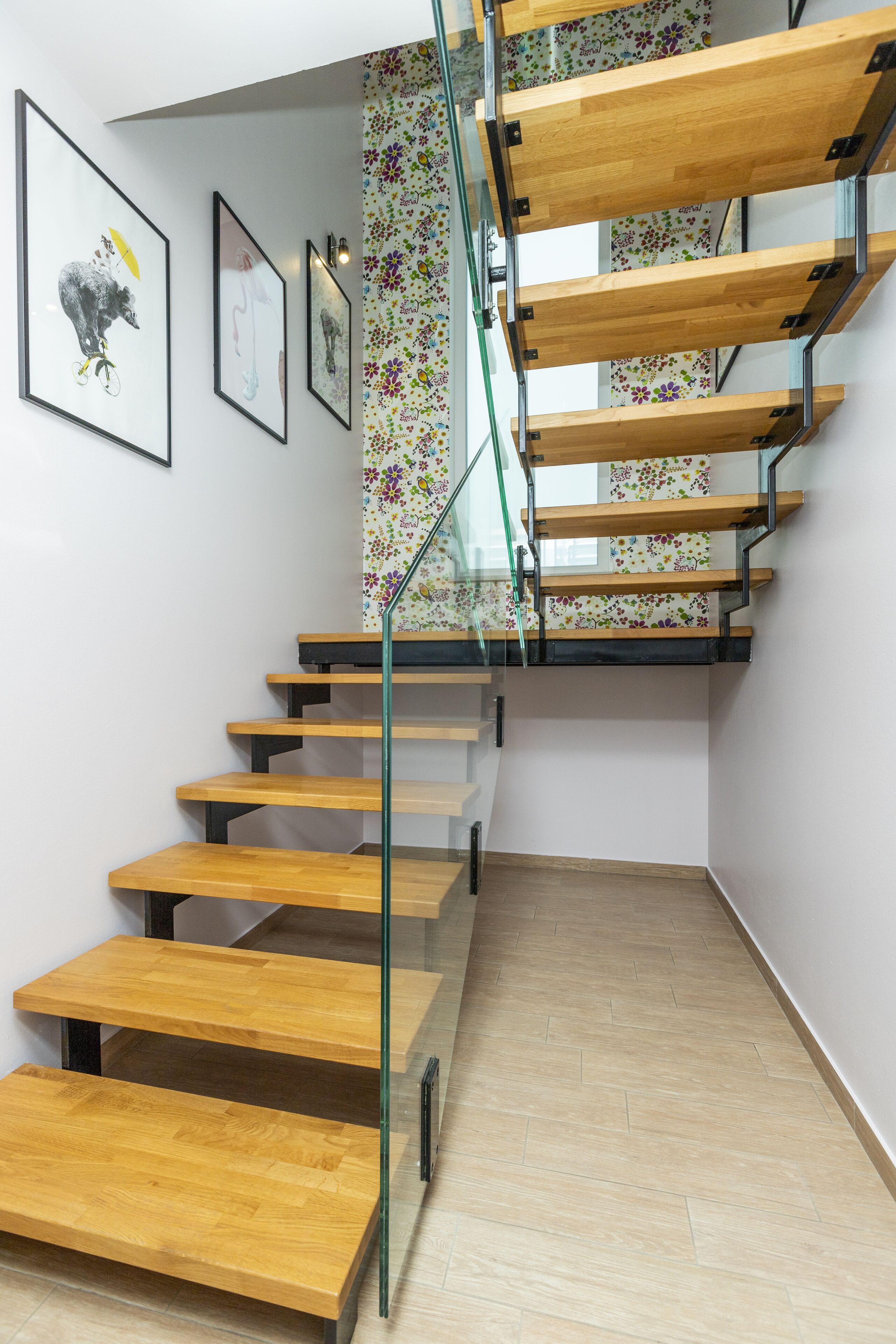 Una escalera moderna con escalones de madera y barandas de vidrio, que conduce a un rellano con un papel tapiz estampado con motivos florales.