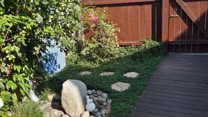 Een houten terras dat leidt naar een tuin met een kiezelpad, een grote rots en een muur met een blauw-witte muurschildering.