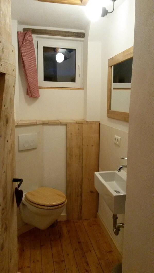A small, compact bathroom with a wooden floor, a toilet, a sink, and a window above the toilet.