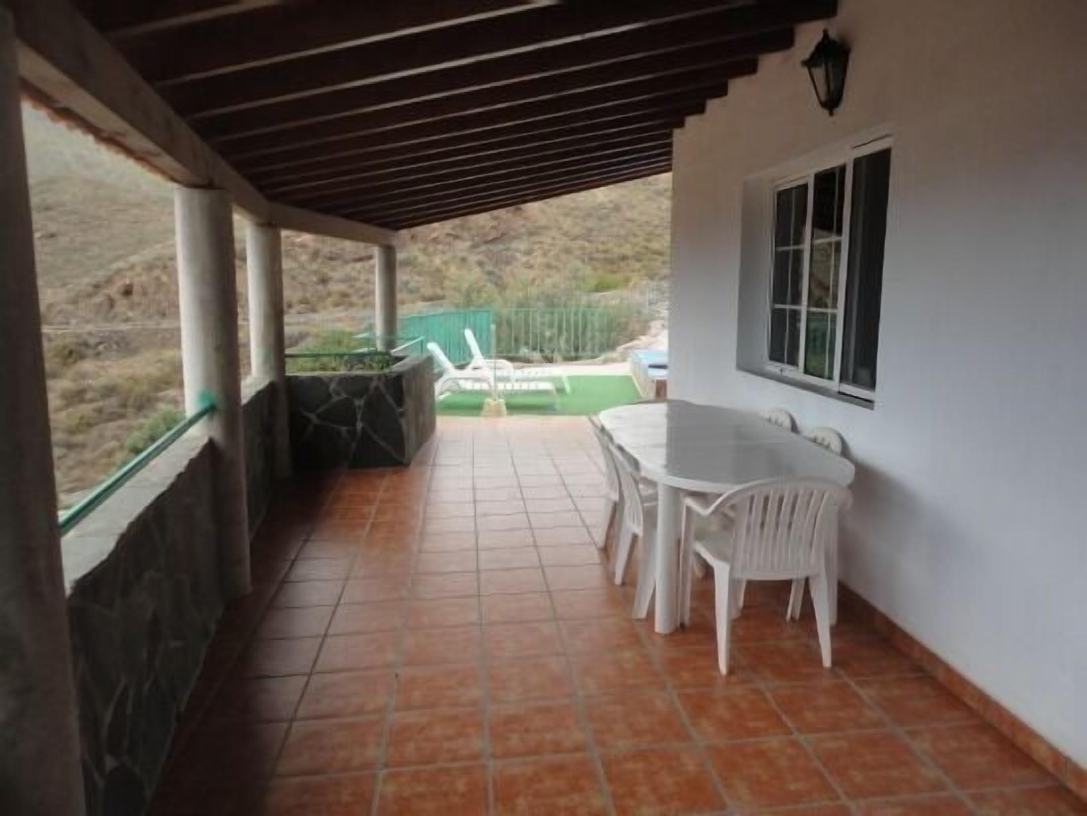 A covered patio with a tiled floor, a white table and chairs, and a window with a view of a green area.