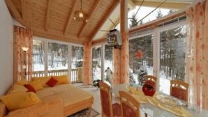 Quarto aconchegante com sofá, mesa de jantar, cadeiras e grandes janelas com vista para uma floresta nevada.
