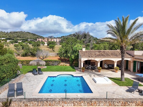 A swimming pool with lounge chairs, a covered patio, and a view of a hillside with buildings.