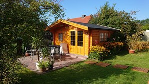 A wooden house with a red roof, a patio with chairs and a table, and a garden with flowers and a lawn.
