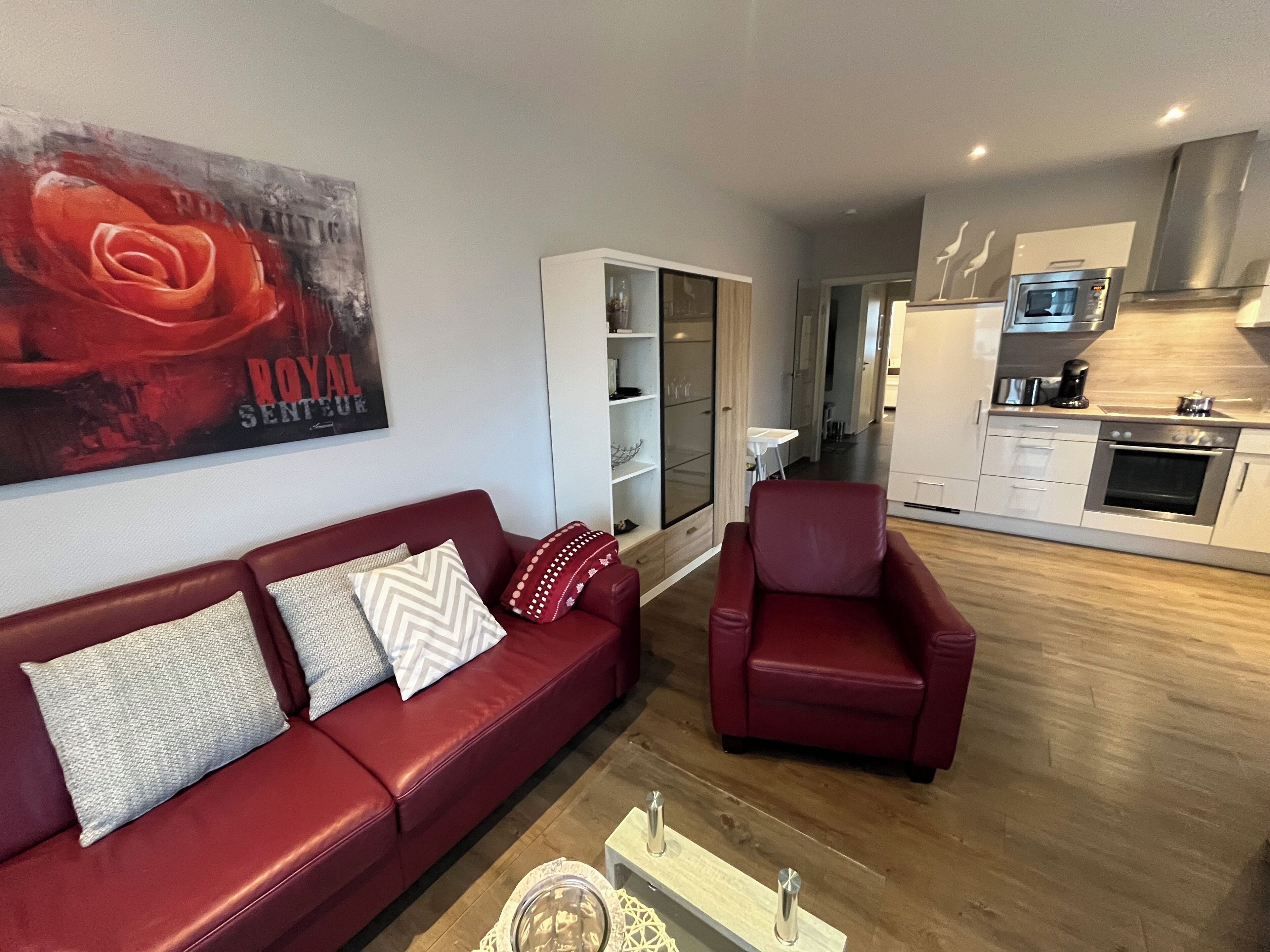 A modern living room with a red sofa and armchair, a glass coffee table, and a kitchen area with stainless steel appliances.
