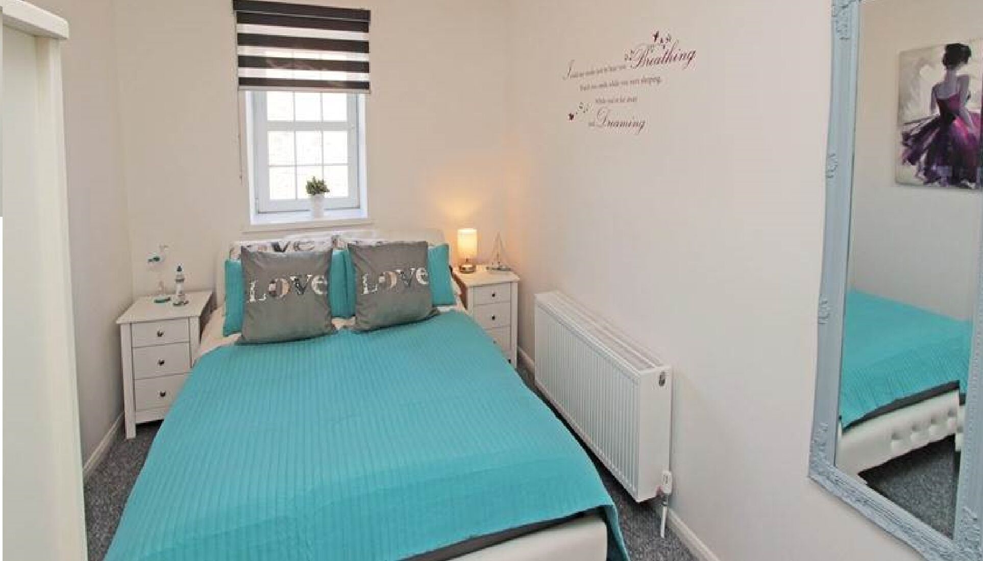 A bedroom with a bed, two bedside tables, a nightstand, a window with blinds, a radiator, and a wall decoration.