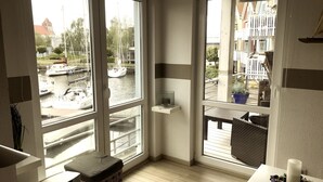 A corner room with large windows overlooking a canal with boats, a small shelf, a bench, and a table with a candle and flowers. - Maritime boutique vacation apartment AM YACHTHAFEN (Greifswald)