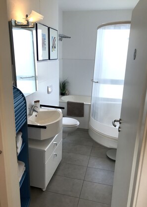 A compact bathroom with a white sink, toilet, and bathtub. There is a towel rack, a small potted plant, and a framed picture on the wall.