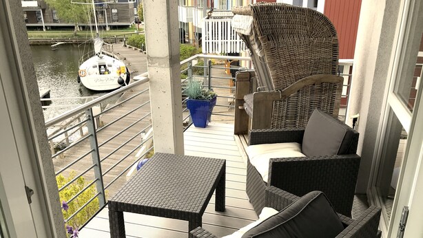 A balcony with wicker furniture, a small table, and a potted plant. A boat is visible in the background. - Maritime boutique vacation apartment AM YACHTHAFEN (Greifswald)
