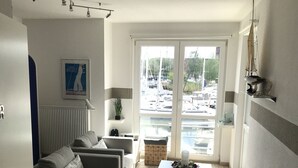 A living room with a sofa, a coffee table, and a view of a marina through large windows. - Maritime boutique vacation apartment AM YACHTHAFEN (Greifswald)