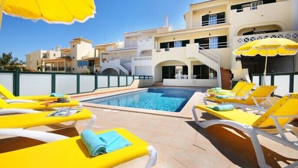 A poolside area with yellow lounge chairs and umbrellas, a clear blue pool, and a building with balconies and a staircase. - 8 bedroom Villa Bella, Private Pool, Albufeira  Close to Strip & Old Town (Albufeira)