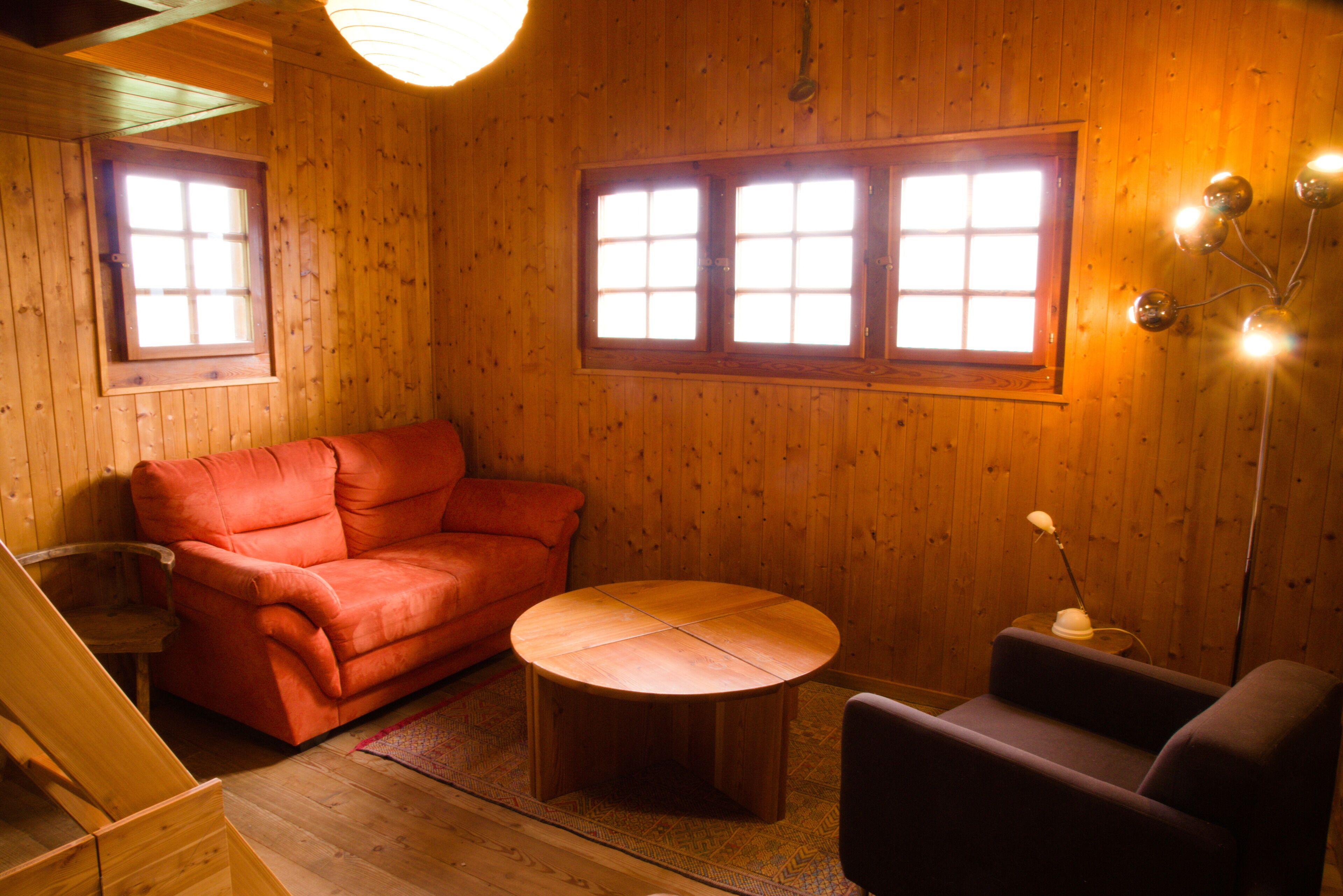 A cozy room with wooden walls, a red sofa, a round wooden table, and two windows providing natural light.