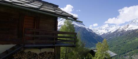 A wooden cabin with a balcony, a pile of firewood, and a mountain backdrop.