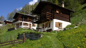 A wooden chalet with a balcony, a stone wall, and a grassy area with yellow flowers. - Magic chalet for 2 people (Villaz/la Sage - Evolène)