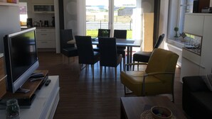 A modern living room with a flat-screen TV, a dining area with chairs, a sofa, and a window with a view of the outdoors. - Vacation home at the Wangermeer, 3 bedrooms up to 8 pers. charging station / wallbox (Hohenkirchen)