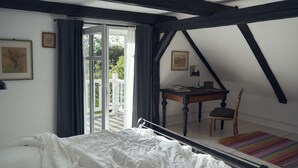 A bedroom with a bed, a desk, and a chair. There is a window with curtains, a door, and a view of the outside.