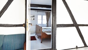 A room with exposed wooden beams, a bed, a window with curtains, and a view of another room.