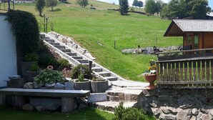 Eine Steintreppe führt zu einer Holzhütte mit Metallgeländer, umgeben von viel Grün und einer Wiese.