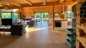 A spacious living area with a sofa, TV, and dining table. There is a kitchen with a refrigerator and cabinets. Large windows allow natural light and views of greenery. - Luxury wooden house with garden on the Baltic Sea with sauna / whirlpool near Swinoujscie (Kolczewo)