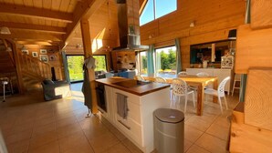 A spacious kitchen with a central island, a dining area with white chairs, and a large window providing a view of the outdoors. - Luxury wooden house with garden on the Baltic Sea with sauna / whirlpool near Swinoujscie (Kolczewo)