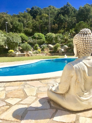 A Buddha statue by a pool with a stone patio, surrounded by lush greenery and a clear blue sky. - A beautiful house surrounded by an idyllic garden with a saltwater pool. (Calonge)