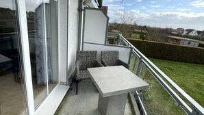 En balkon med et betonbord og fletstole, en skydedør af glas og udsigt til et boligområde.