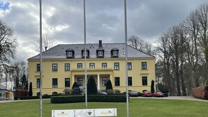 En gul bygning med et tag af fliser, omgivet af en velplejet græsplæne og træer.