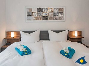A double bed with white linens, two pillows, and a blue towel with flowers. There are two bedside tables with lamps. A wall art piece is above the bed.