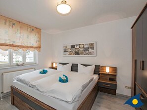 A bedroom with a double bed, two bedside tables, a nightstand, a wall-mounted artwork, and a window with a curtain. - Vacation apartment Strandläufer - Whg. 06 // - Strandläufer 06 (Heringsdorf)