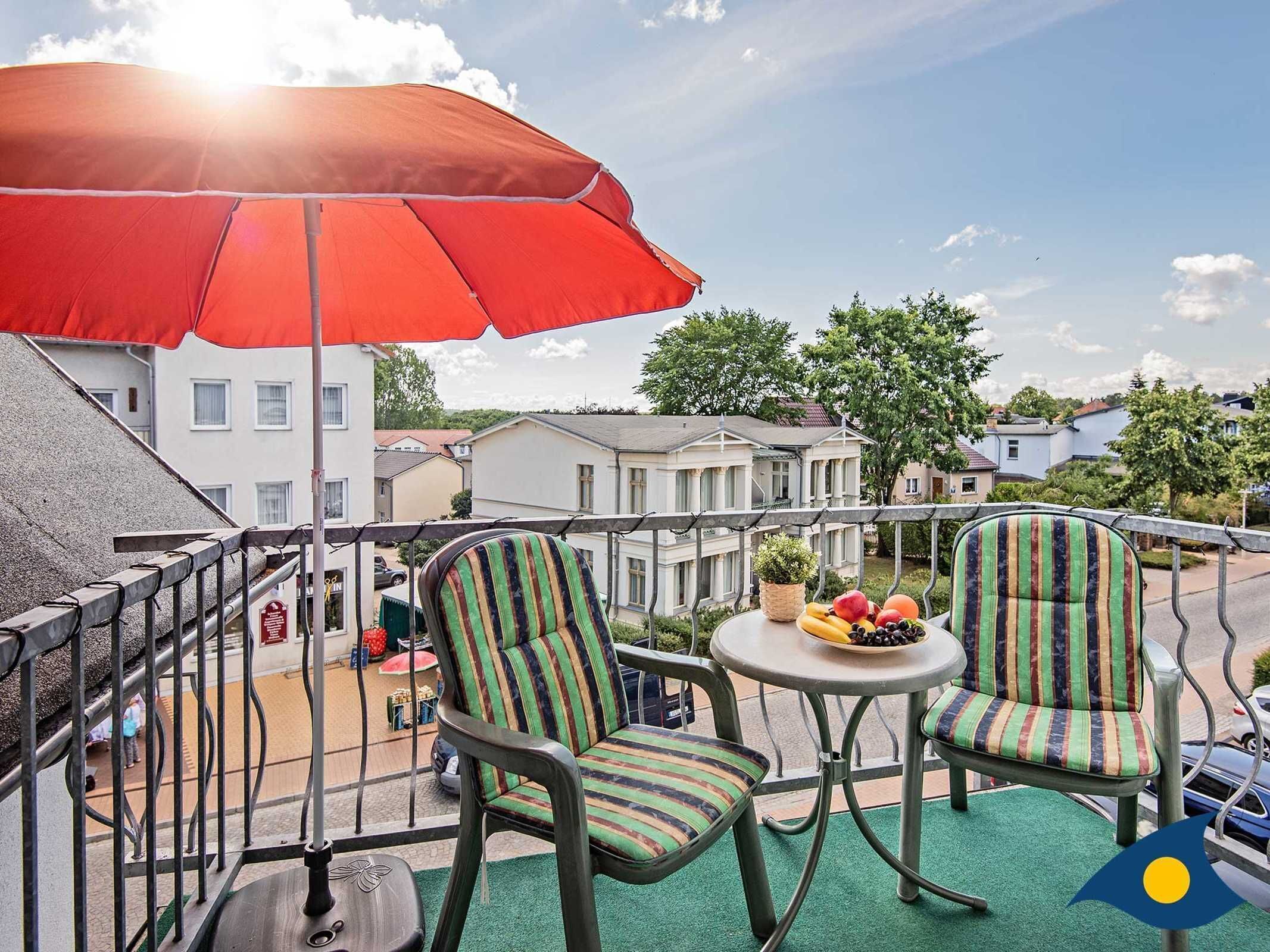 Un balcon avec deux chaises à rayures, une petite table ronde sur laquelle se trouvent des fruits et un parasol rouge.