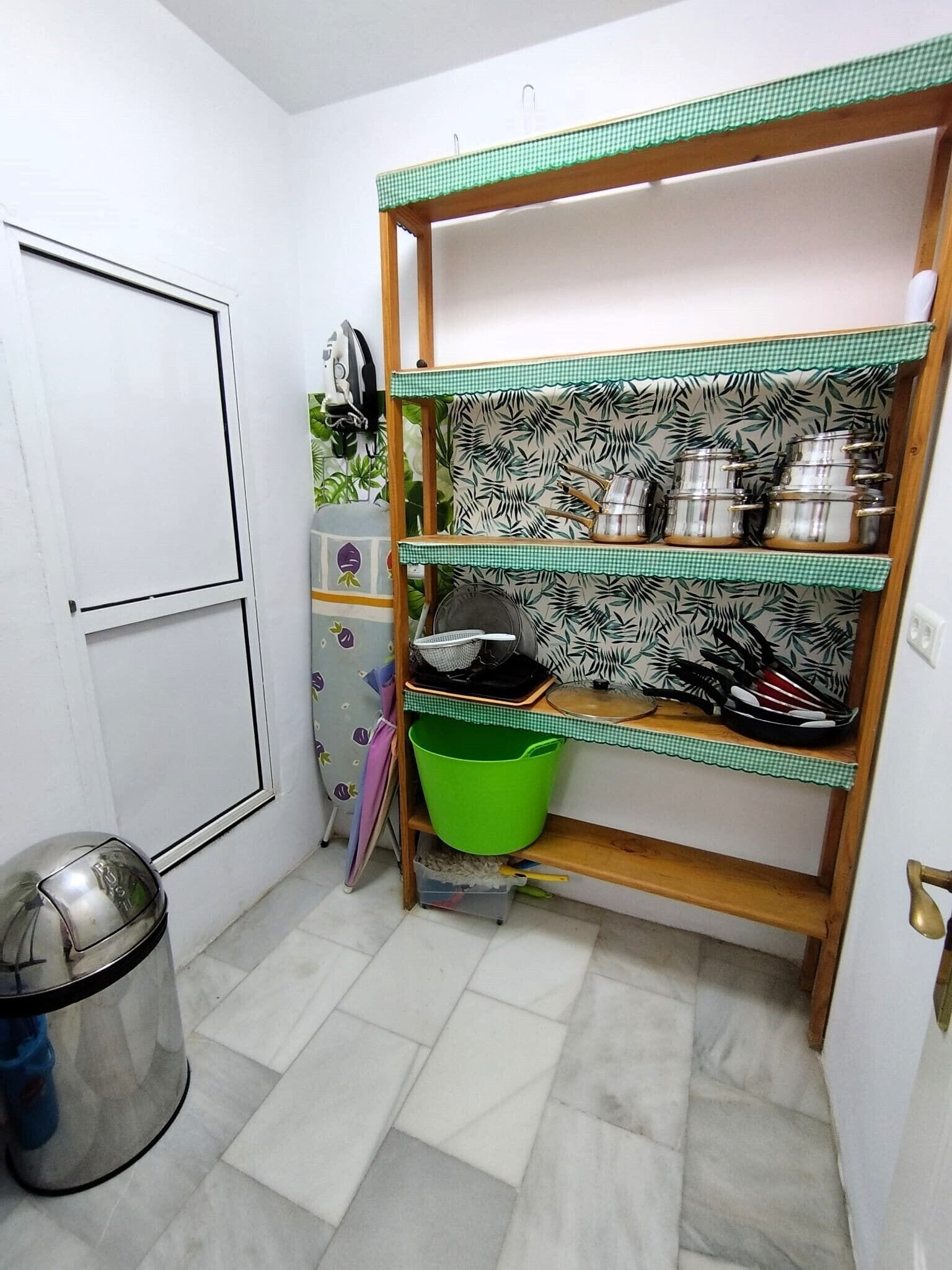 A compact kitchen with a white door, a green trash bin, and shelves with kitchenware.