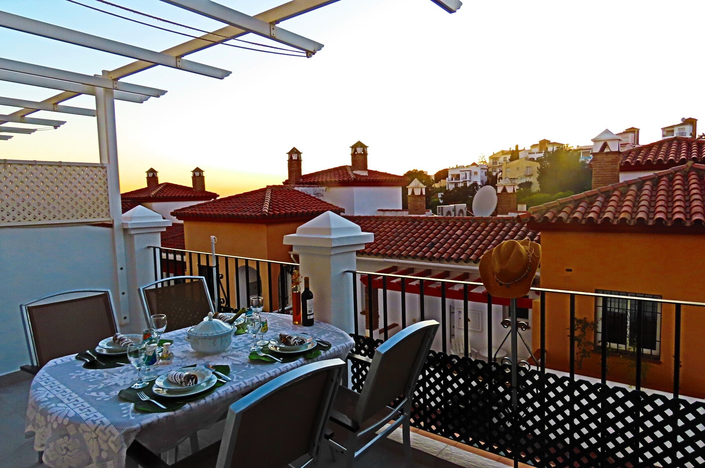 A balcony with a table set for dining, overlooking a residential area with red-tiled roofs and white walls.