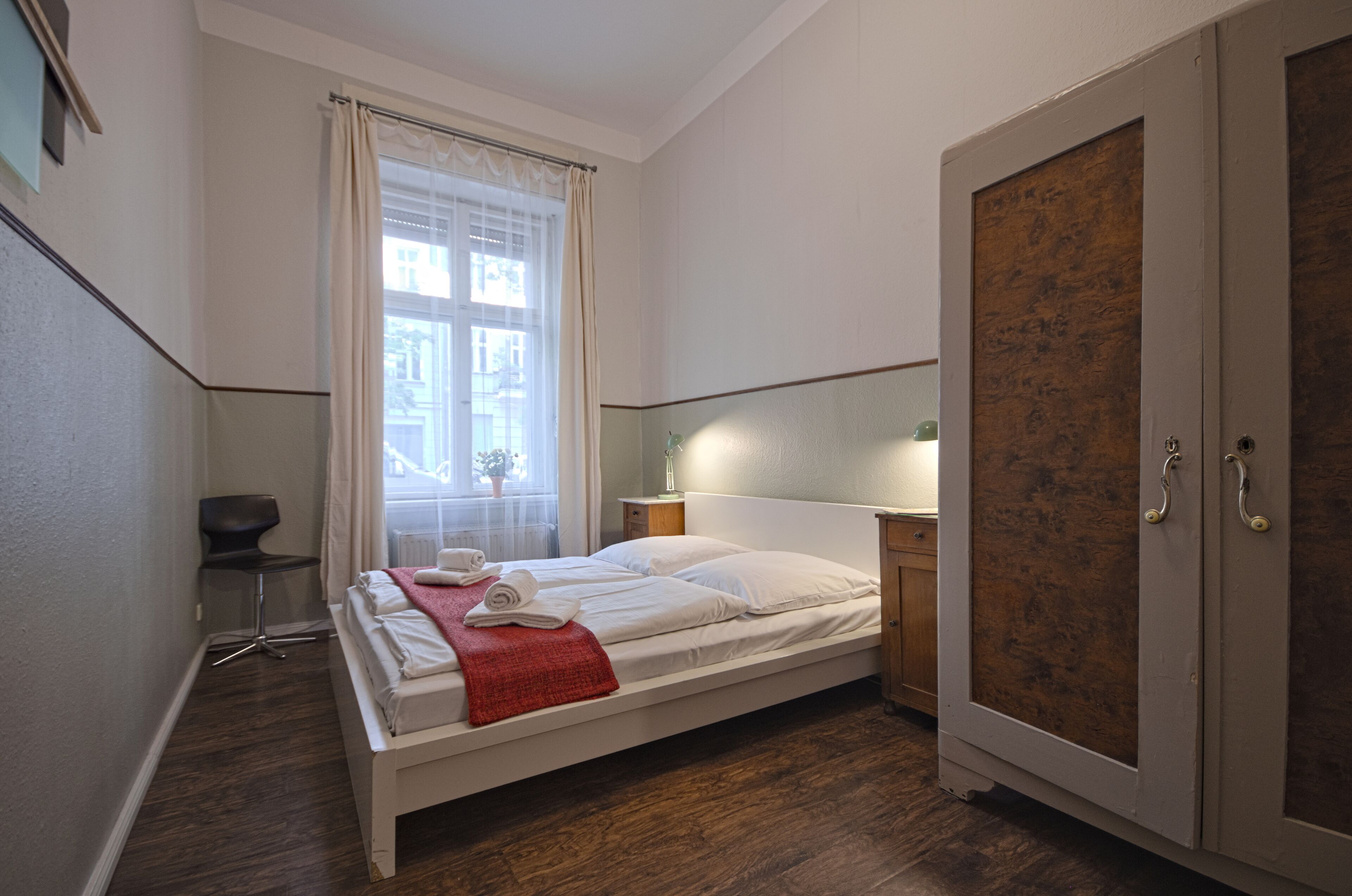 Een net opgemaakt bed met witte lakens, een rode plaid en een stoel ernaast. Er is een raam met dunne gordijnen en een lamp op een nachtkastje.