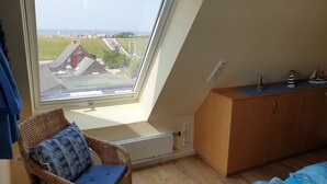 A room with a large skylight, a wicker chair with a cushion, a wooden cabinet, and a view of a building and greenery outside. - Cozy attic studio Norddeich panoramic window with sea view beach 50 meters (Norden)