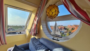 A room with a large window, a round skylight, a model sailboat, and a blue sofa. - Cozy attic studio Norddeich panoramic window with sea view beach 50 meters (Norden)