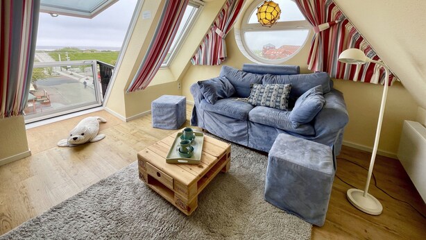 A cozy loft bedroom with a large skylight, a blue sofa, a wooden coffee table, and a fluffy white dog. - Cozy attic studio Norddeich panoramic window with sea view beach 50 meters (Norden)