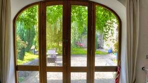 Une vue à travers une porte en bois dotée d’un panneau de verre, qui donne sur un espace extérieur avec un chemin pavé, une table et des chaises.